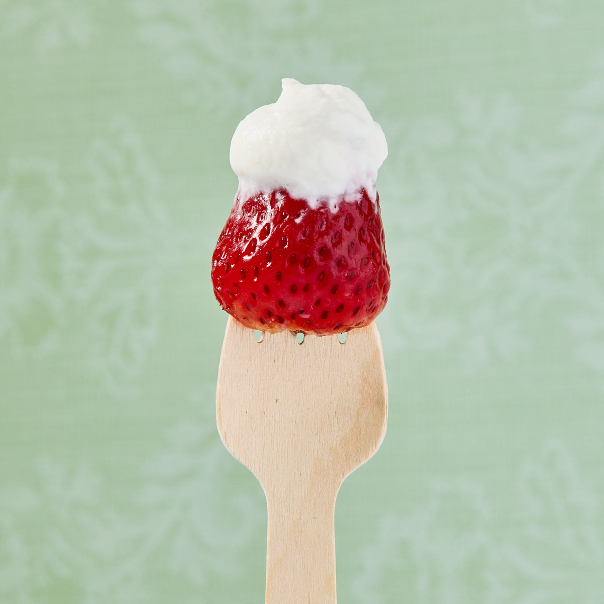 A close-up of a strawberry topped with whipped white cream on a wooden birchwood spoon by Birchware, set against a pale green patterned background.