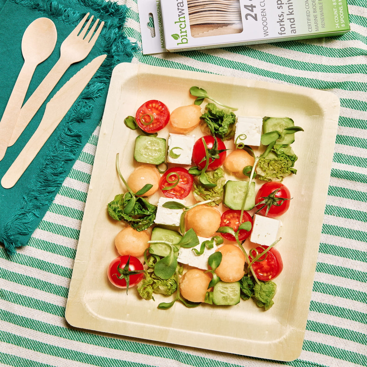 A styled flat-lay of a set of 24 wooden forks, knives, and spoons by Birchware shown in their packaging at the top, alongside three natural wood utensils on a teal napkin and a green and white striped tablecloth. The utensils are arranged on a disposable and compostable wooden serving platter with a colorful salad.
