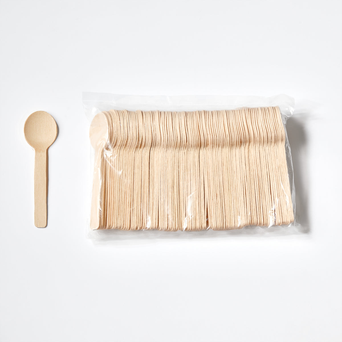 A pack of light natural wood disposable and compostable birchwood mini spoons in a clear plastic bag, shown next to a single matching wooden spoon on a clean white background.